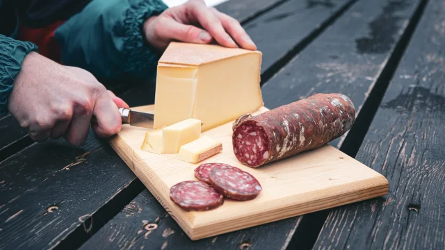 Une planche en bois sert de support à un saucisson pré-découpé tandis que quelqu'un coupe des tranches d'Abondance avec un Opinel.