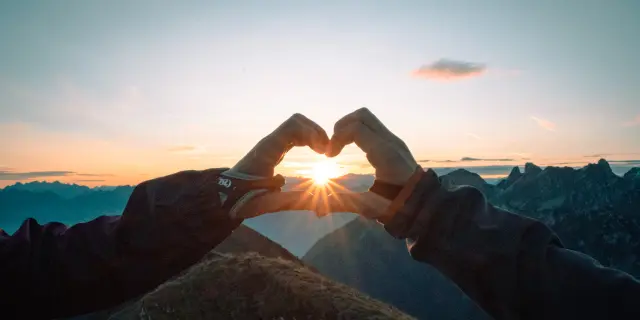 Un coeur fait avec les doigts, entourant le soleil couchant, face aux montagnes.