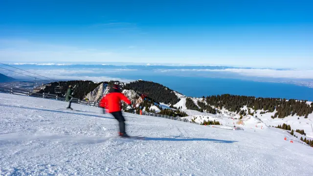 Un skieur en train de descendre une piste, sur les hauteurs de Thollon-les-Mémises, avec vue sur les montagnes suisses, le Léman, ainsi qu'un grand ciel bleu et ensoleillé